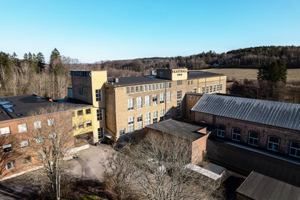 Fabriksbyggnad i tegel med skyltarna 'KASTHALL' och 'MALTFABRIK', omgiven av träd och åkrar under klarblå himmel.
