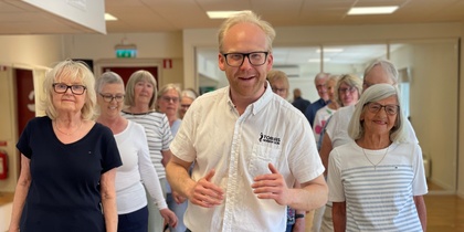 En grupp äldre personer och en yngre man står tillsammans inomhus, leende och avslappnade.