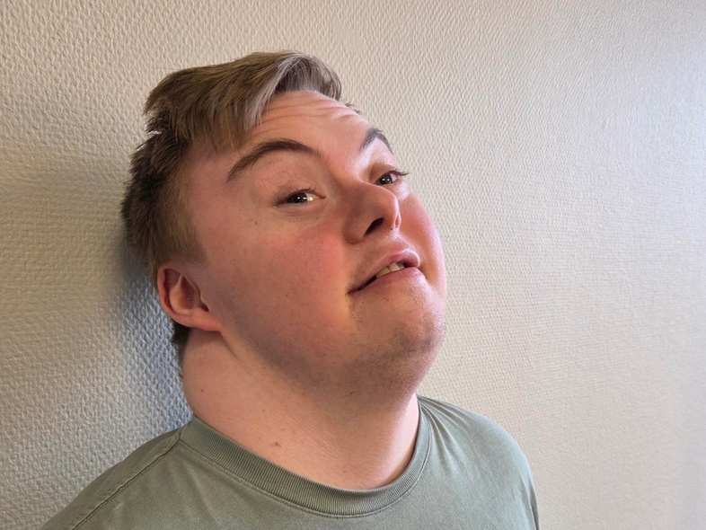 Person i grön t-shirt står lutad mot en ljus vägg med huvudet bakåtlutat. Ansiktet är suddat.