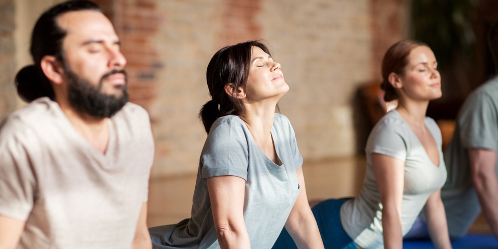 Tre personer gör en yogaövning på blå mattor inomhus, med ansiktena vända uppåt och ögonen slutna.