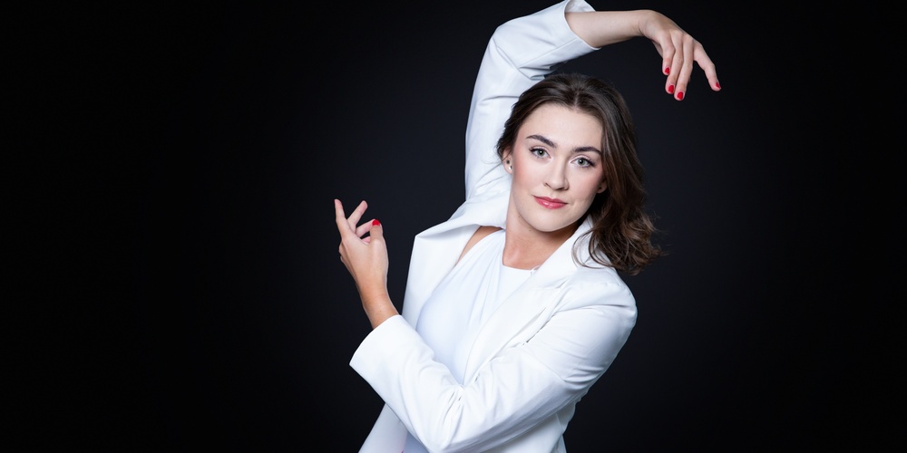 Person i vit kavaj och rosa byxor poserar konstnärligt mot svart bakgrund.