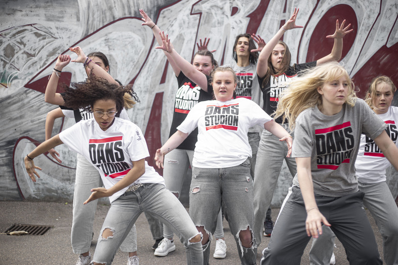 En grupp unga kvinnor dansar energiskt utomhus framför en graffitivägg, alla bär t-shirts med texten 'DANS STUDIO'.