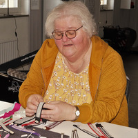 En person sitter vid ett bord och arbetar med band och textilier i en ljus inomhusmiljö.