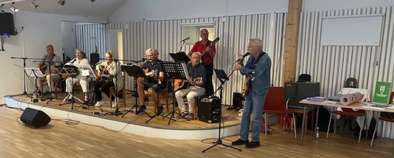 Ett band med äldre personer spelar musik på en liten scen i ett ljust rum med trädetaljer.