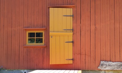 En röd trävägg med ett litet gult fönster och en gul dörr ovanför marknivå.