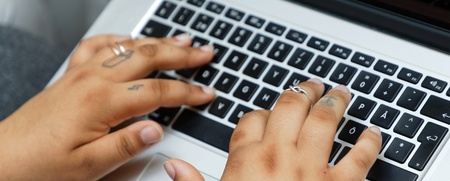 Två händer med ringar och tatueringar skriver på ett svartvitt tangentbord på en bärbar dator.