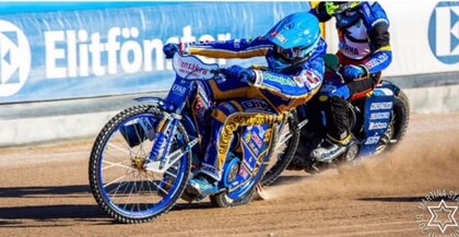 Två motorcykelförare tävlar på en grusbana, båda i färgglada dräkter och hjälmar.