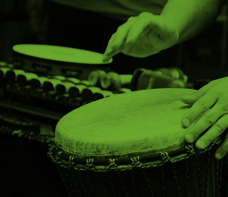 Två händer spelar på en trumma, med fler slagverksinstrument i bakgrunden. Bilden har grön ton.