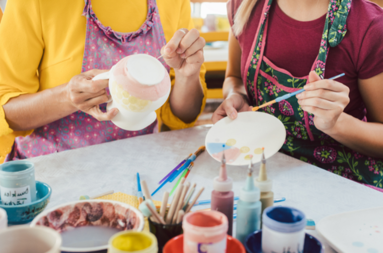 Två personer målar keramikföremål vid ett bord fyllt med färger och penslar.