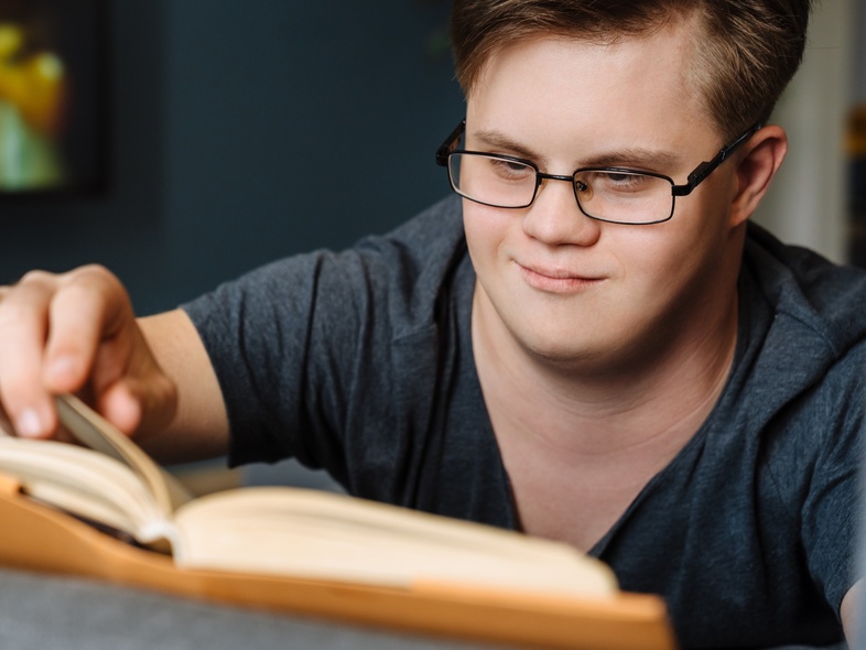 Ung man med glasögon och Downs syndrom läser en bok och vänder sida i ett vardagsrum.