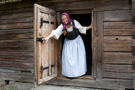 Kvinna i traditionella kläder står i dörröppningen till en gammal timmerstuga och ler.