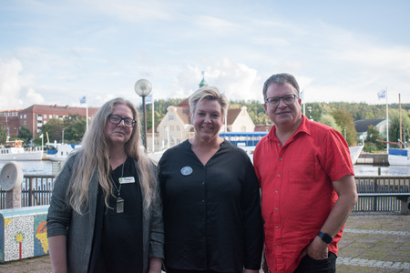 Tre personer står utomhus vid en hamn med båtar och byggnader i bakgrunden.