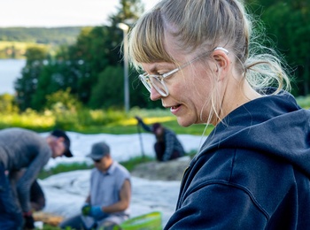 En kvinna med glasögon och uppsatt hår arbetar på en odlingsmark med andra personer i bakgrunden.