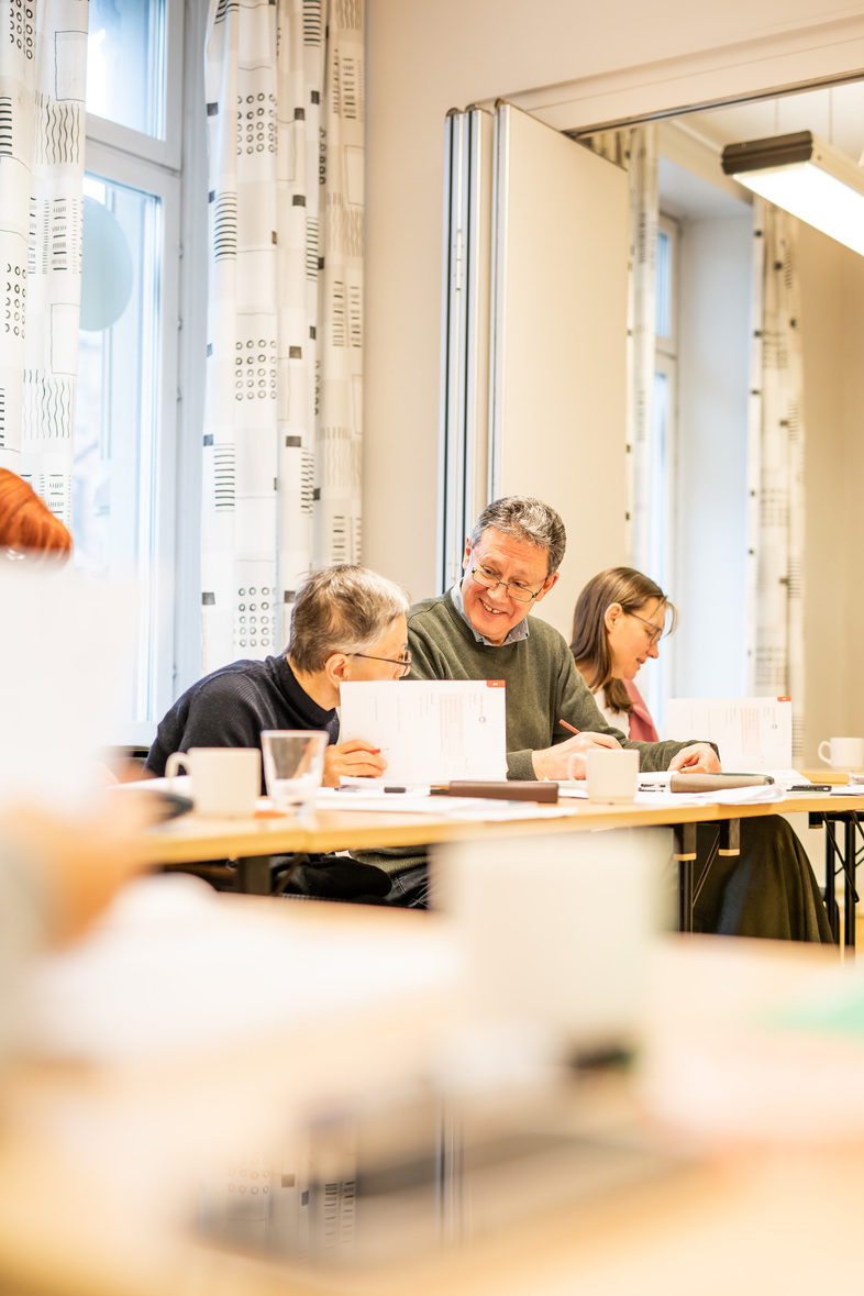 Tre personer sitter vid ett bord i ett ljust rum och samtalar, med papper och kaffekoppar framför sig.