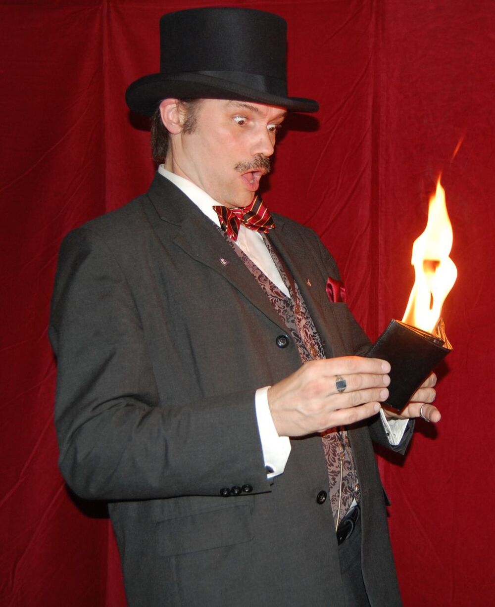 Man i kostym och hög hatt håller en plånbok med eldslåga, ser förvånad ut mot röd bakgrund.