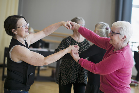 Två äldre kvinnor dansar tillsammans i ett ljust rum, leende och håller varandras händer.