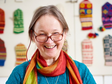 Leende kvinna med glasögon, blå tröja och färgglad halsduk mot en suddig bakgrund med stickade vantar i olika traditionella mönster på väggen bakom henne