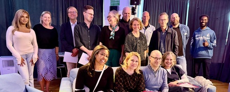 Gruppbild med 15 personer, några sittande i soffor och andra stående, i ett rum med blåa och svarta gardiner.