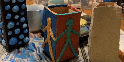 Tre färgglada, handgjorda pappersfigurer står på ett bord med konstmaterial och färg i bakgrunden.