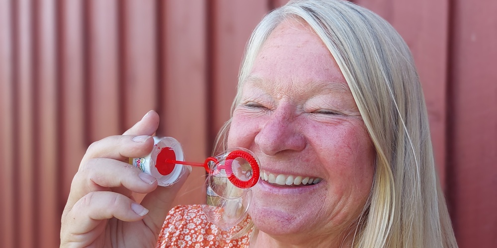 Leende kvinna blåser såpbubblor framför en röd trävägg.