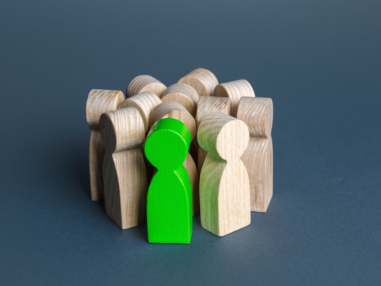 Träfigurer står i en grupp, en av dem är grön medan resten är träfärgade, mot en blå bakgrund.