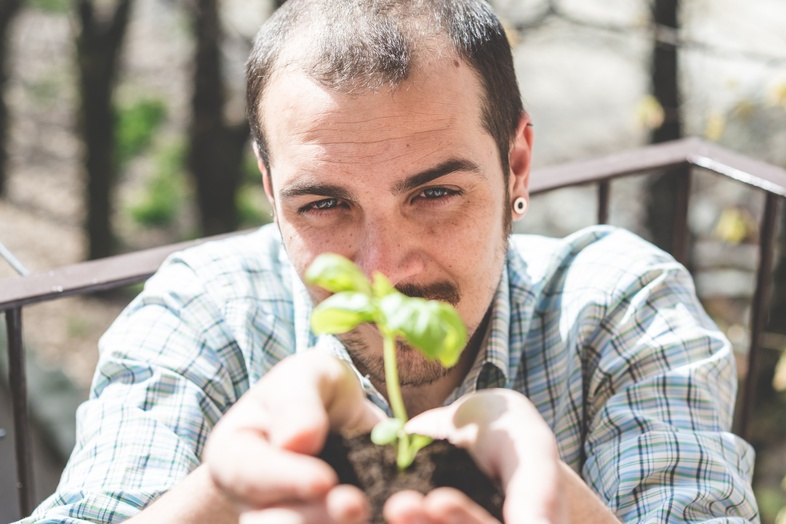 En man håller en liten planta med jord i händerna och ler mot kameran utomhus.
