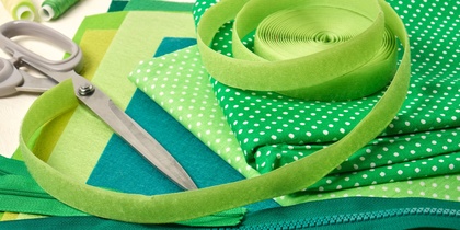 Sytillbehör i gröna nyanser, inklusive tyg, sax, dragkedja, tråd och band, ligger utspridda på ett bord.