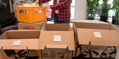 Person sorterar saker i flyttlådor märkta med slänga, sälja och skänka i ett ljust rum.