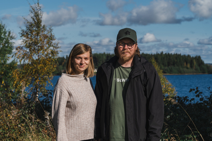 En kvinna och en man står utomhus vid en sjö med skog i bakgrunden under en solig dag.