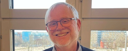 En person står inomhus framför stora fönster med utsikt över en stadsmiljö och parkerade bilar.