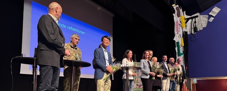 En grupp personer står på en scen med mikrofoner och blommor, framför en projicerad presentation.