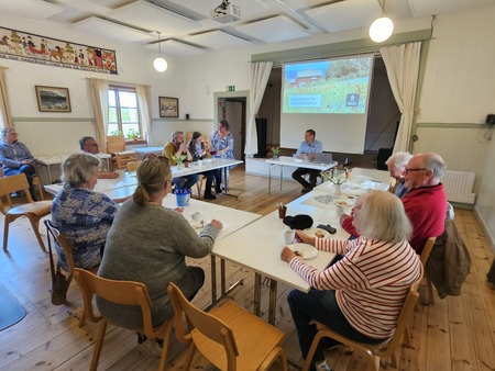 En grupp äldre personer sitter runt bord i ett ljust rum och deltar i en presentation med projektor.