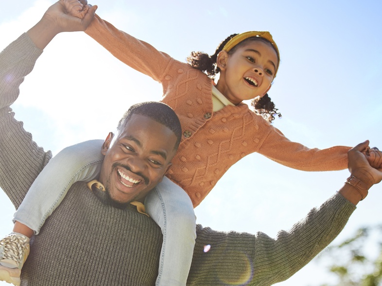 En man bär en leende flicka på sina axlar utomhus under en solig dag.