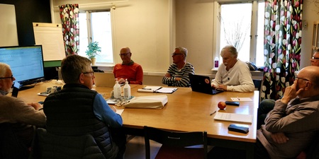 Sju personer sitter runt ett bord i ett mötesrum med datorer, papper och en stor skärm.