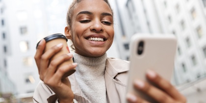 Ung kvinna ler och tittar på sin mobiltelefon utomhus, håller en kaffekopp i andra handen.