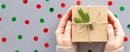 Två händer håller ett inslaget paket med snöre och grankvist mot en bakgrund med röda och gröna prickar.