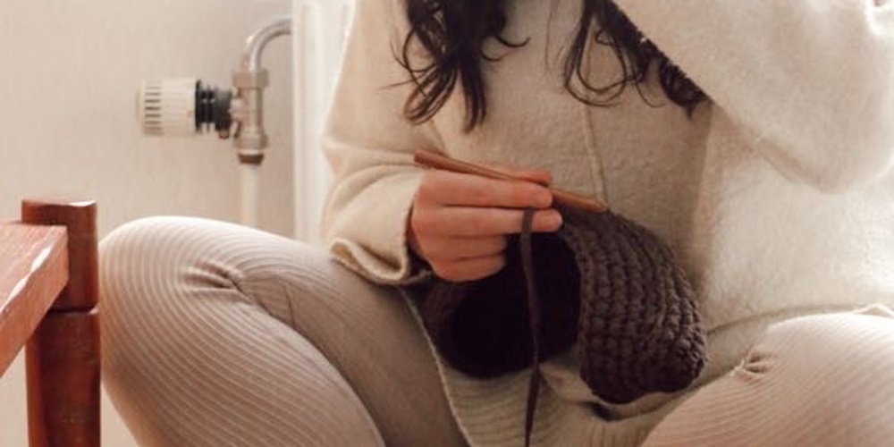 Person sitter korslagd på en kudde och stickar med mörkbrunt garn i ett ljust, mysigt rum.