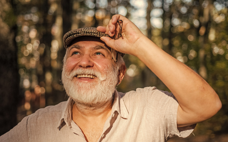 Leende äldre man med vitt skägg och keps står utomhus i soligt skogsparti.