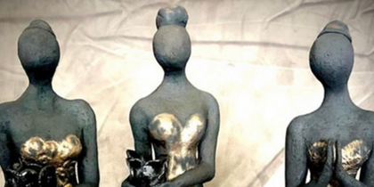 Tre stiliserade kvinnliga statyetter i långa klänningar, utan ansikten, står på rad mot en neutral bakgrund.