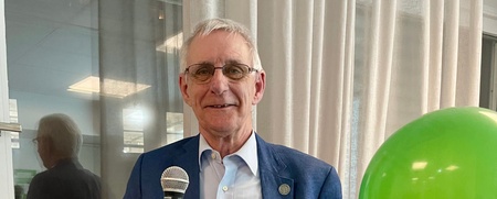 Äldre man i blå kavaj håller mikrofon och presentpåse, står bredvid ett grönt ballong vid ett fönster.