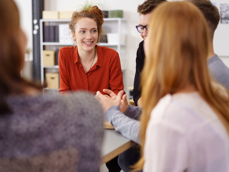 Fyra personer sitter runt ett bord och samtalar, en kvinna i röd skjorta ler mot gruppen.