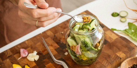 En person lägger grönsaker i en glasburk på en skärbräda, omgiven av ingredienser för inläggning.