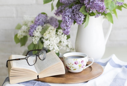 En öppen bok med glasögon, en kopp te och en vas med lila och vita blommor på ett bord.