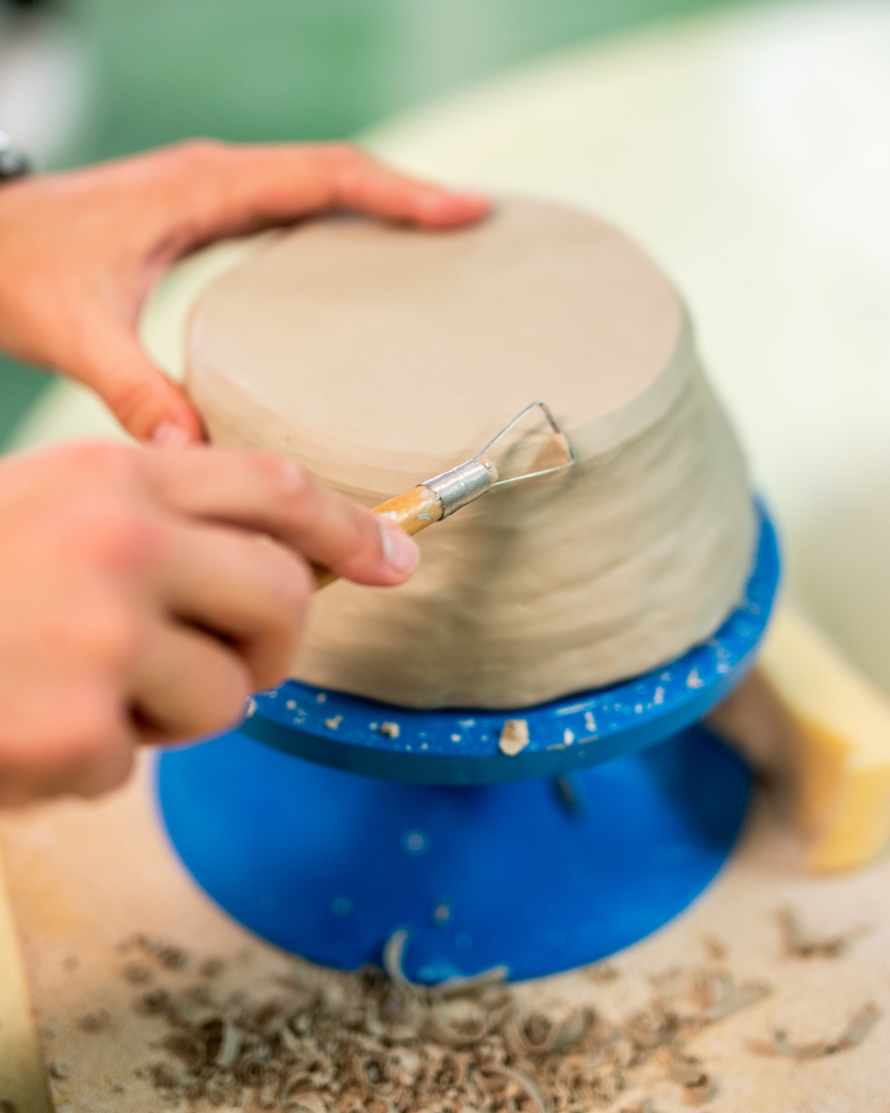 En person beskär en lerkruka med ett verktyg på en blå drejskiva.