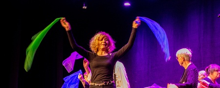 En grupp människor på scen viftar med färgglada tygsjalar under en föreställning med lila scenbelysning.
