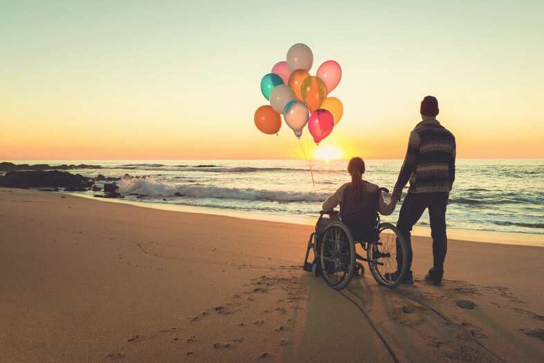 Två personer tittar på en solnedgång på en sandstrand. Den ena personen sitter i en rullstol och håller en knippe ballonger i olika färger i ena handen.