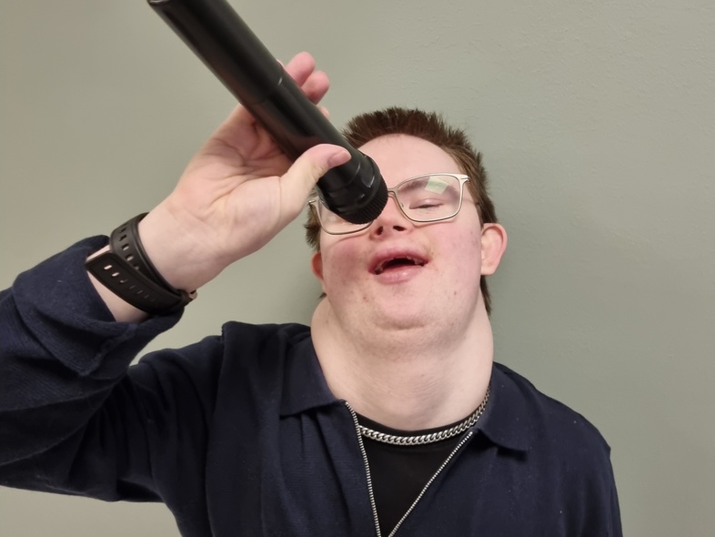 Person håller ett svart cylinderformat föremål mot huvudet, ansiktet är suddat, bakgrund i ljusgrå färg.