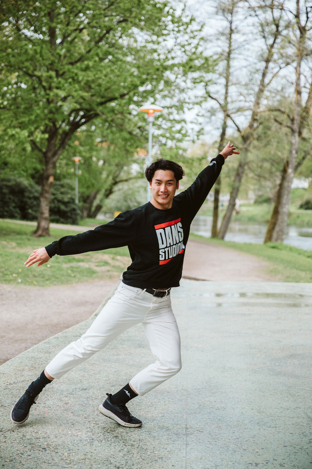 En ung man dansar utomhus på en gångväg, omgiven av gröna träd och gräs.