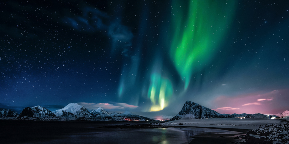 Norrsken lyser upp natthimlen över snötäckta berg och en frusen kust.