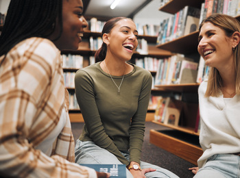 Tre unga kvinnor sitter och skrattar tillsammans i ett bibliotek.